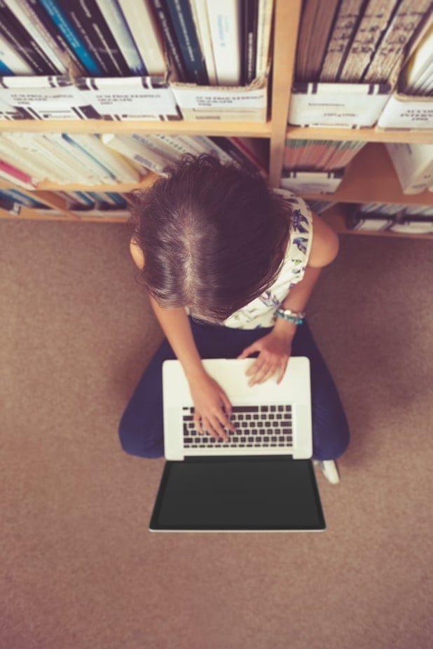 High angle view of a female student sitting against bookshelf and using laptop on the library floor.jpeg